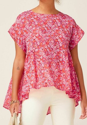 Femme portant un haut fleuri rose à manches courtes avec un ourlet asymétrique et un pantalon écru, tenant un sac tissé beige dans sa main gauche.