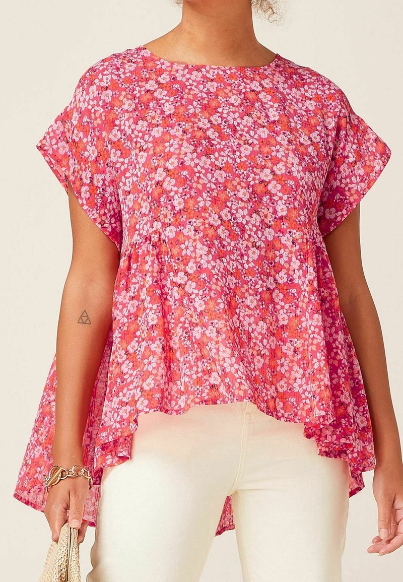 Femme portant un haut fleuri rose à manches courtes avec un ourlet asymétrique et un pantalon écru, tenant un sac tissé beige dans sa main gauche.