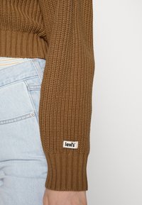 Manche de pull tricoté marron avec étiquette logo Levi's, portée avec un jean en denim bleu clair sur fond blanc uni.