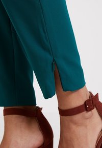 Teal colored trousers with a slit at the hem, paired with brown ankle strap heels. Fabric appears smooth with a slight sheen.