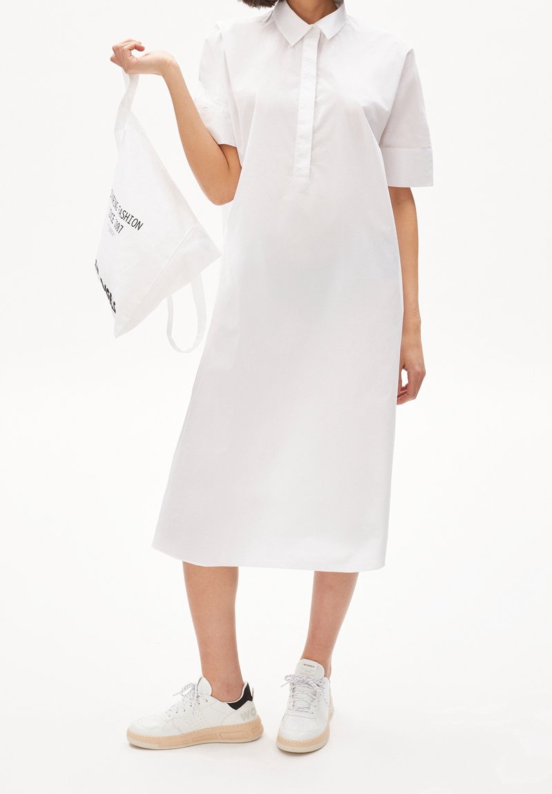 Personne en robe chemise blanche et baskets blanches tenant un tote bag blanc avec du texte noir sur un fond blanc uni.