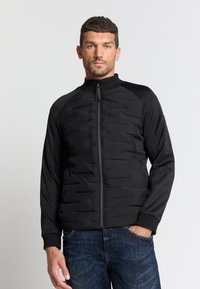 Zwarte pufferjack met een rits aan de voorkant, geribbelde kraag en een gladde stof. Beschikt over gewatteerde details op het lichaam en zachte mouwen.