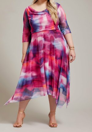 Kobieta ubrana w kolorową, różową, fioletową i niebieską sukienkę midi z efektem tie-dye, z rękawami do połowy przedramienia oraz beżowe sandały na wysokim obcasie, stojąca na jednolitym tle.