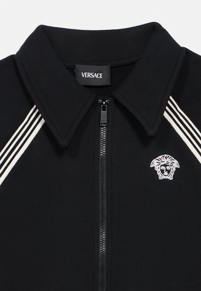 Μαύρο τζάκετ με φερμουάρ και γιακά πόλο, λευκές ρίγες και λευκό κεντητό λογότυπο Medusa στο στήθος. Ύφασμα με μείγμα βαμβακιού.