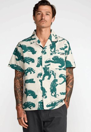 Hombre con tatuajes que lleva una camisa de manga corta con estampados de panteras verdes y un bolsillo en el pecho sobre un fondo claro.