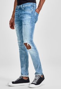 Light blue denim jeans with distressed details, featuring a ripped knee, slim fit, five pockets, and paired with black sneakers.