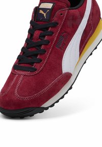Adidași roșii din velur cu o dungi albă Puma, șireturi negre, accent galben pe talpă și talpă exterioară din cauciuc texturat negru.
