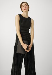 Svart ermeløs topp med subtil perledetalj, parret med svarte, plisserte vidbenede bukser. Modellen holder en svart clutch med en duskdetalj.