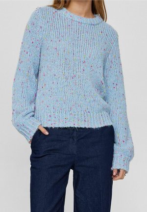 Femme portant un pull en grosse maille bleu clair avec des taches multicolores et un pantalon bleu foncé, main dans la poche.