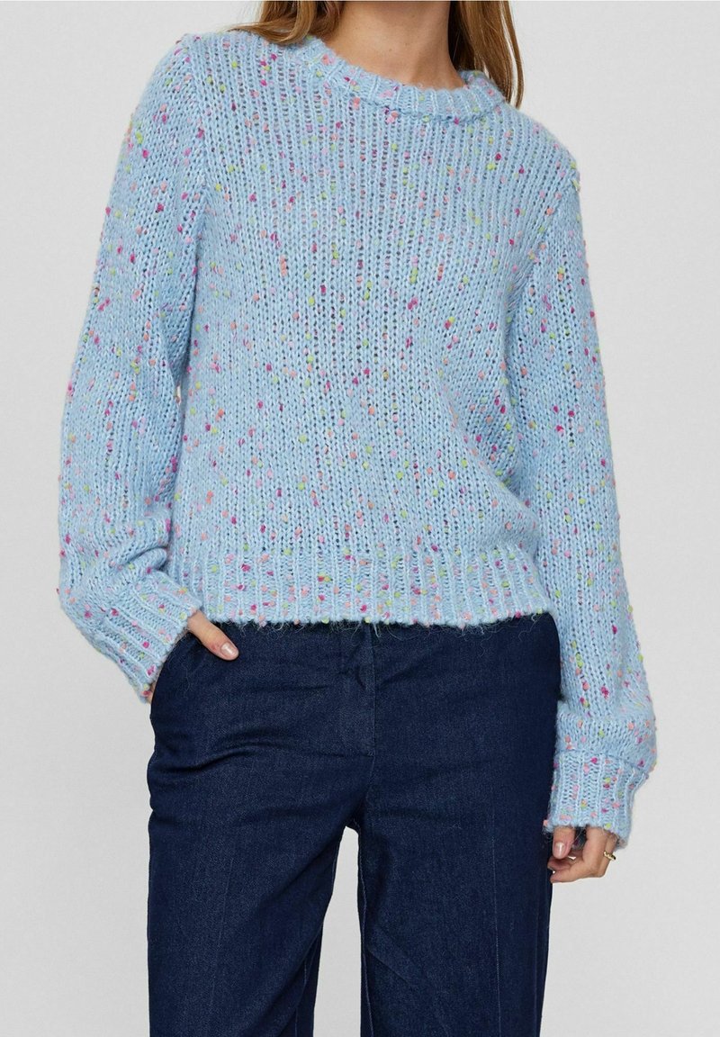 Femme portant un pull en grosse maille bleu clair avec des taches multicolores et un pantalon bleu foncé, main dans la poche.