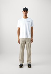 T-shirt bianco in cotone con testo ricamato, pantaloni cargo beige con tasche laterali e sneakers nere con suola a contrasto.