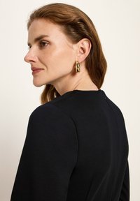 Frau mit hellbraunem Haar, die ein schwarzes Top und goldene Creolen trägt, sieht nach links vor einem schlichten hellen Hintergrund.