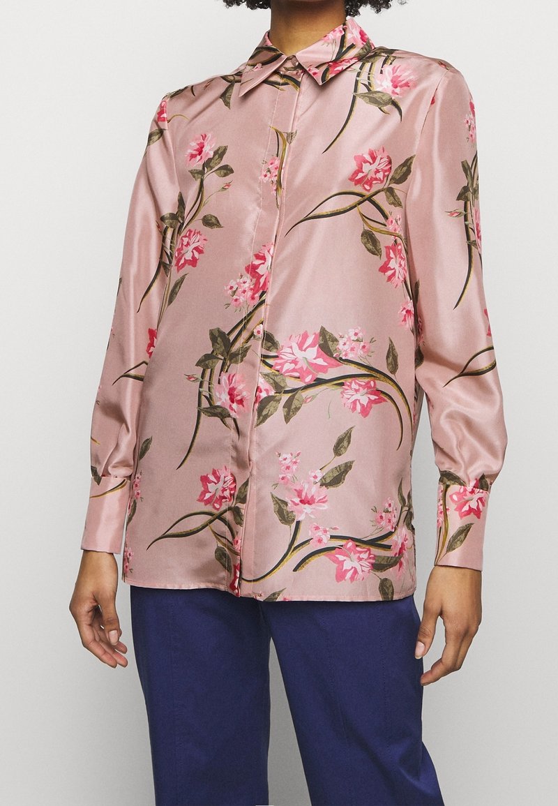 Personne portant une chemise rose satinée à manches longues avec des imprimés floraux et un pantalon bleu, debout devant un fond blanc uni.