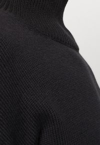 Pull à col roulé noir en tricot avec un motif de points texturé, montrant la zone de l'épaule et du cou contre un fond clair uni.