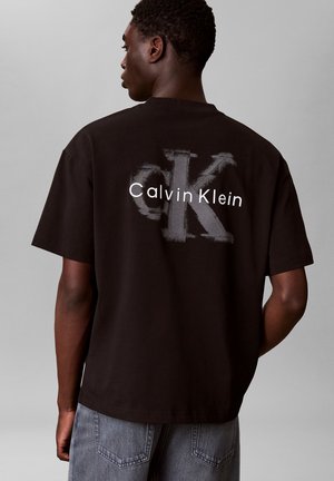 Junger Mann trägt ein schwarzes Calvin Klein T-Shirt mit großem, abgenutztem "CK"-Logo und Aufschrift "Calvin Klein" auf dem Rücken.