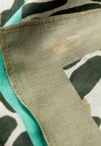 Bomuldsstof med et grønt camouflage mønster og en kontrasterende turkis kant. Synlig syning supplerer teksturens udseende.
