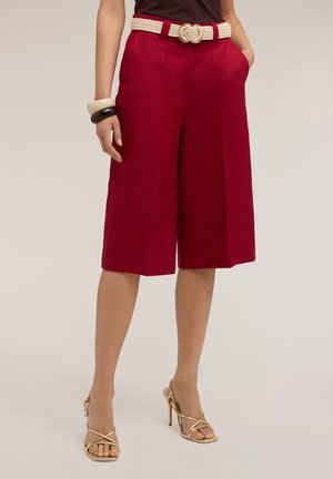 Donna che indossa pantaloncini culotte rossi con cintura intrecciata bianca, top nero, sandali dorati con tacco alto e braccialetti spessi al polso sinistro.