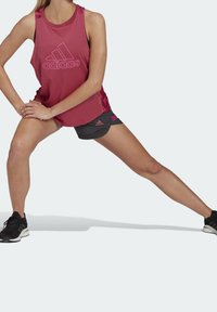 Czerwona sportowa koszulka na ramiączkach z dużym różowym logo Adidas, zestawiona z ciemnoszarymi szortami. Modelka w pozycji wykroku, ubrana w czarne buty sportowe.