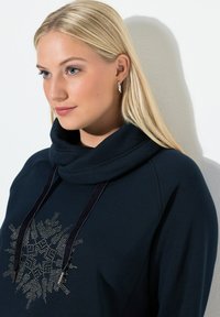 Sudadera con capucha de color azul marino y cuello tipo cowl, con un patrón de copos de nieve brillantes. Tiene cordones oscuros y una textura suave.