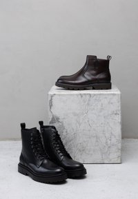 Czarne skórzane botki na sznurowadła z teksturowanymi podeszwami oraz brązowe skórzane botki wsuwane z pętelką do zakładania, prezentowane na marmurowym piedestale.