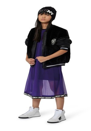 Chaqueta bomber de piel sintética negra sobre un vestido transparente morado, combinada con zapatillas altas blancas y un gorro negro con logo.