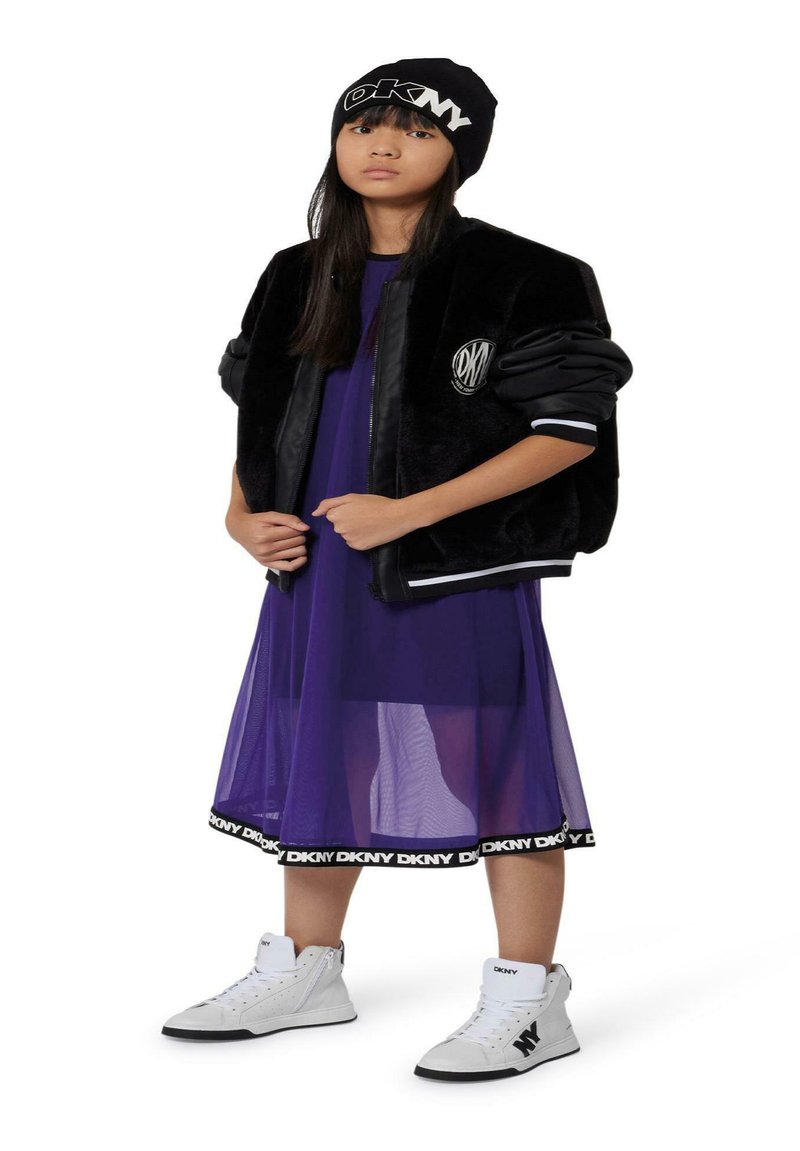 Veste bomber en fausse fourrure noire sur une robe transparente violette, assortie à des baskets montantes blanches et un bonnet noir avec logo.
