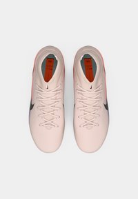 Ζευγάρι ανοιχτό ροζ ποδοσφαιρικών παπουτσιών Nike με μαύρα λογότυπα swoosh, λευκά κορδόνια και πορτοκαλί πάτους, που απεικονίζονται από πάνω σε λευκή επιφάνεια.