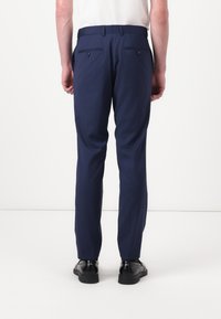 Marineblaue maßgeschneiderte Hosen mit glatter Textur, slim Fit und doppelt genähten Gesäßtaschen, kombiniert mit einem Gürtel und getragen mit schwarzen Schuhen.