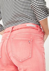 Jeans de mezclilla rosa con un borde deshilachado en la cintura, que cuentan con un pequeño bordado de un flamenco blanco en el bolsillo trasero. Camisa de cuadros visible arriba.