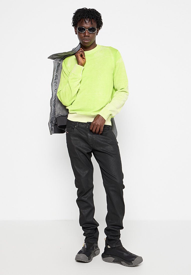 Helle limonengrüne Sweater in einem taillierten Design, kombiniert mit schwarzen Jeans und schwarzen Sportschuhen. Das Model hält eine graue Jacke über der Schulter.