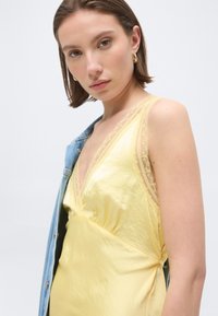 Νεαρή γυναίκα με κοντά καστανά μαλλιά φοράει χρυσούς κρίκους στα αυτιά, κίτρινο φόρεμα με δαντέλα και ένα τζιν μπουφάν κρεμασμένο στον έναν ώμο.