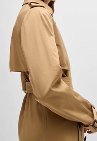 Widok z boku osoby ubranej w beżowy płaszcz trench z paskiem w talii i klapą na ramieniu na jednolitym tle.