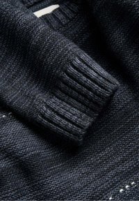 Dunkel gestrickter Pullover mit einem strukturierten Stoff, gerippten Bündchen und einem subtilen Muster. Das Material wirkt weich und dick.