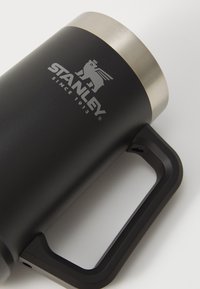 Musta ruostumattomasta teräksestä valmistettu matkakuppi, jossa on kaareva muoto, hopeinen vuoristorata ylhäällä ja musta kahva, jossa on Stanleyn logo.