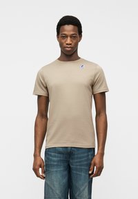 Άνδρας που φοράει μπεζ κοντομάνικη μπλούζα με μικρό λογότυπο στο στήθος και μπλε τζιν, που στέκεται μπροστά σε απλό φόντο.