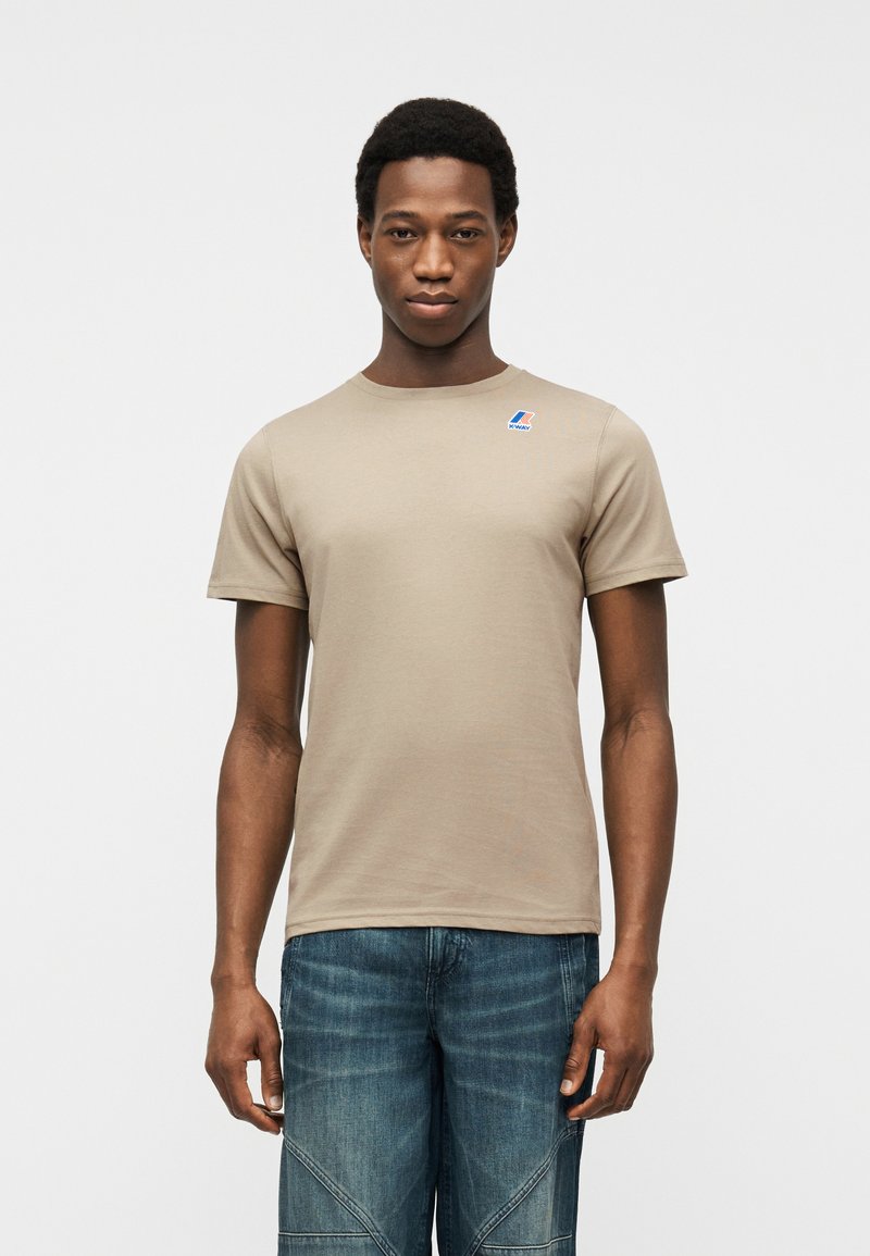 Άνδρας που φοράει μπεζ κοντομάνικη μπλούζα με μικρό λογότυπο στο στήθος και μπλε τζιν, που στέκεται μπροστά σε απλό φόντο.