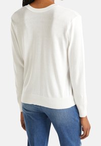 Vit stickad tröja med rund halsringning, ribbade ärmslut och nederkant. Slät textur med en något lös passform, bärs över blå jeans.