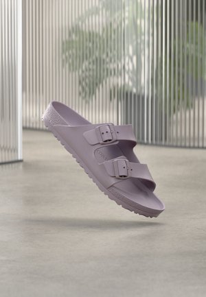 Egy lavendula színű papucs két állítható csatos pánttal, texturált talppal, sima padlón, a levegőben bemutatva, elmosódott függőleges redőnyös háttérrel.