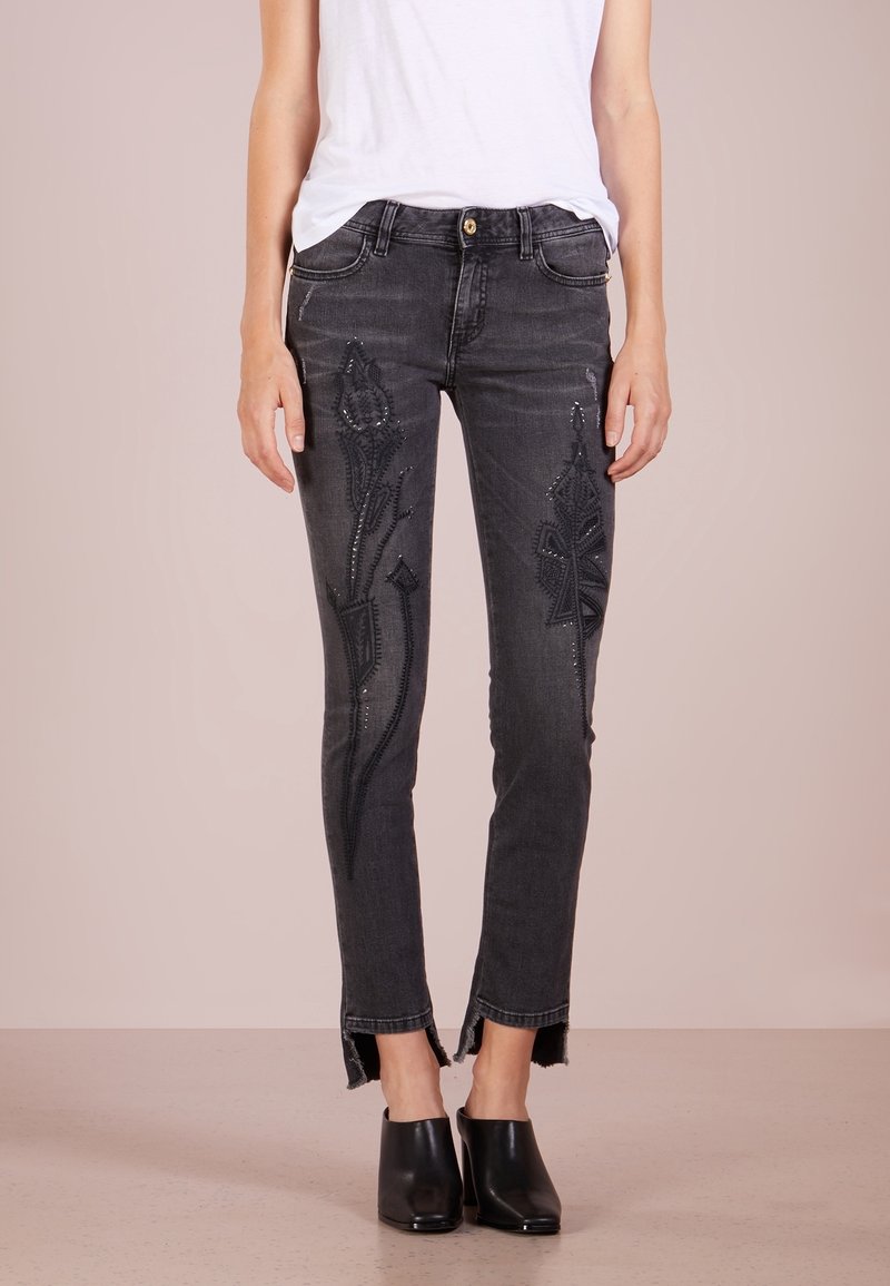 Mujer que lleva unos pantalones skinny gris oscuro con bordados florales negros en los muslos y mulas negras de tacón, de pie contra un fondo liso.