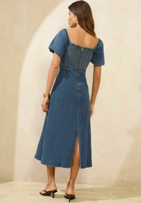 Denim midi obleka s smučarskim hrbtom, kratkimi puf rukavi, zadrgo na sredini hrbta in stranskim razporkom. Ima strukturirano prileganje in gladko teksturo.