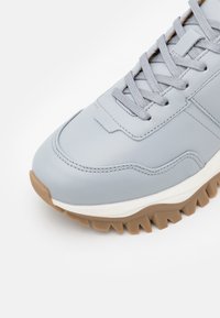 Světlomodré kožené tenisky s texturovaným povrchem, šedými tkaničkami a bílou mezipodešví. Mají robustní podešev z gumy s výrazným vzorem.