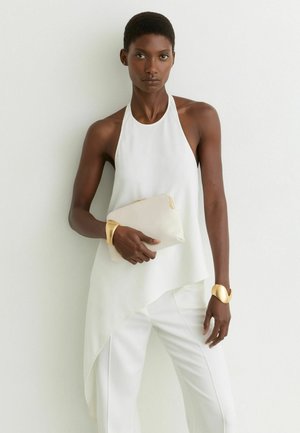 Donna con top bianco asimmetrico senza maniche e pantaloni bianchi, che tiene una pochette beige, con polsini dorati su entrambi i polsi.