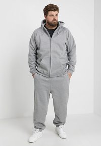 Sudadera gris con cremallera y pantalones de chándal a juego, elaborados en una tela suave, con un corte relajado y bolsillos frontales. Unos tenis blancos completan el conjunto.