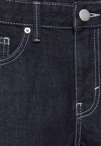 Jean en denim bleu foncé à l'avant avec des coutures blanches, des rivets en argent et un bouton en métal gravé avec le texte de la marque.