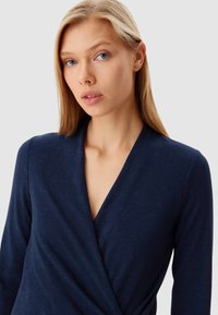Top avvolgente blu navy realizzato in tessuto strutturato, con scollo a V profondo e maniche lunghe, arricchito da sottili accenti scintillanti su tutta la superficie.