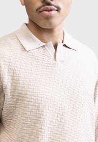 Suéter bege texturizado com decote em colarinho e padrão de diamante. O tecido parece suave, com um brilho subtil e um design leve.