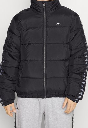 Personne portant une doudoune noire avec des patches de logo blancs sur les manches et la poitrine, associée à un pantalon de jogging gris clair avec une bande de logo noire.