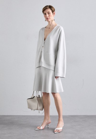 Ανοιχτό γκρι υπερμεγέθη cardigan με V-λαιμόκοψη, κουμπί κλεισίματος και δύο τσέπες, συνδυασμένο με μια ταιριαστή φούστα με φάρδος και μινιμαλιστικά λευκά τακούνια.