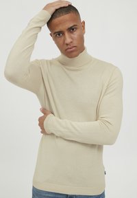 Bež turtleneck pulover iz fine pletene tkanine, ki ima dolge rokave in rebrasto obrobo na ovratniku in zapestjih.
