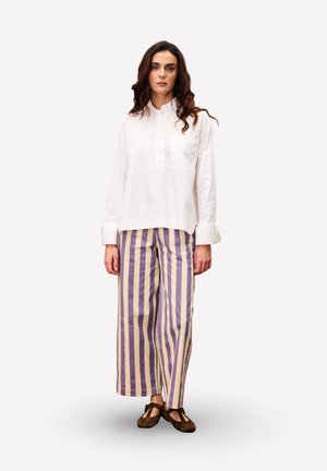 Witte langemouwen shirt met een placket en zak, gecombineerd met brede, gestreepte broek in paars en beige. Bruine schoenen met veters.